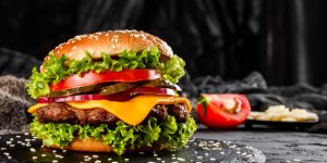Beef burger with cheese, tomatoes, red onions, cucumber and lettuce on black slate over dark background. Unhealthy food